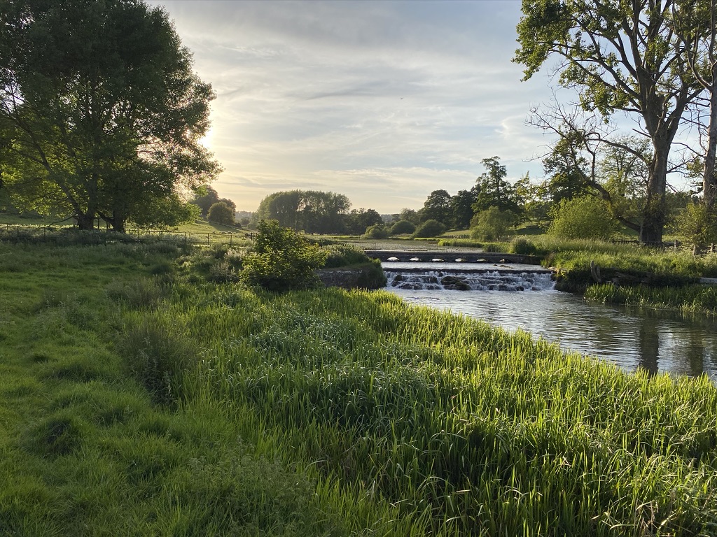 Sherborne Brook flowing over a low weir through a green riverside landscape